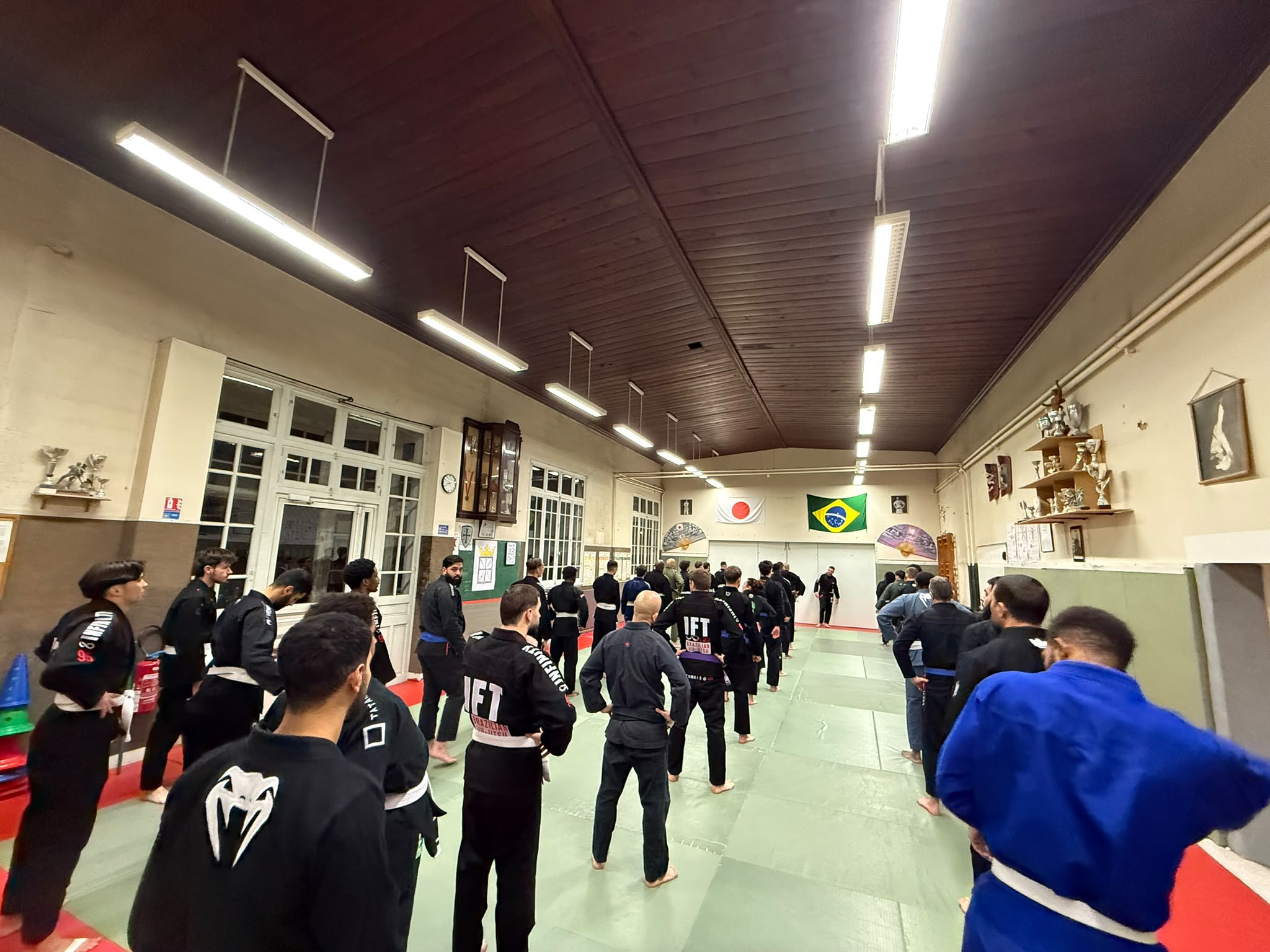 Vue large du dojo pendant un cours collectif, pratiquants rassemblés sur le tatami sous les drapeaux
